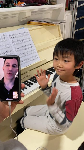 young boy at piano waving while facetiming with piano teacher