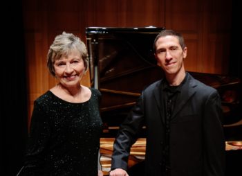 Woman and man (both in black) stand posed in front of a piano