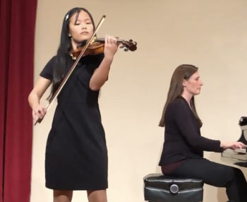 Young woman in black playing violin with a female pianist behind her