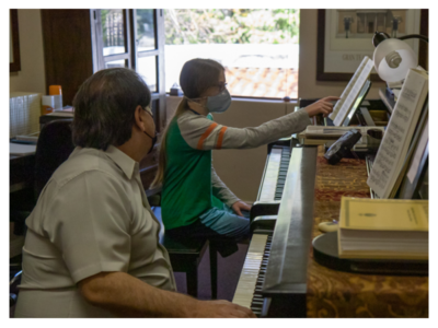 Man with face mask at one piano instructs boy at second piano who is pointing at sheet music