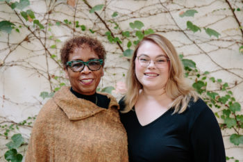 Two women with glasses posing happily in front of a wall of ivy.