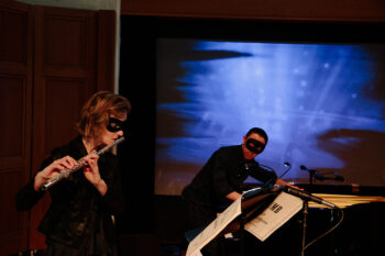 A man playing piano and a woman playing flute, both are wearing eye masks