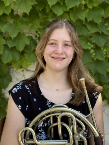 Young woman holding a French horn poses in front of a wall of ivy