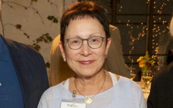 A woman with short hair, a blue shirt, and glasses, smiles